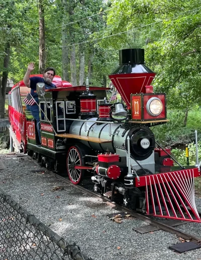 Red miniature train traveling through the woods at Burlington City Park.